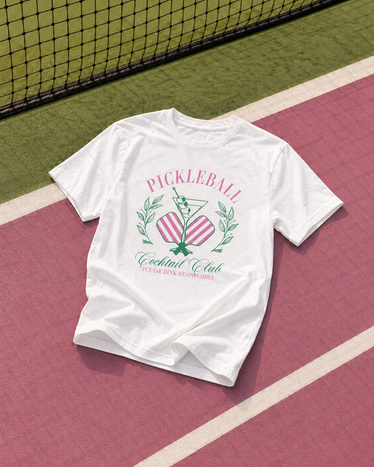 T-shirt blanc imprimé Pickleball Cocktail Club posé à plat sur un terrain de sport rose et vert près du filet.