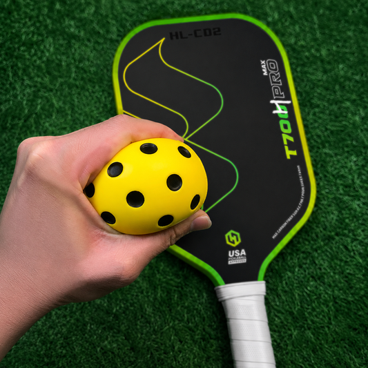Main pressant une balle en mousse jaune silencieuse et flexible devant une raquette de pickleball sur fond de pelouse.