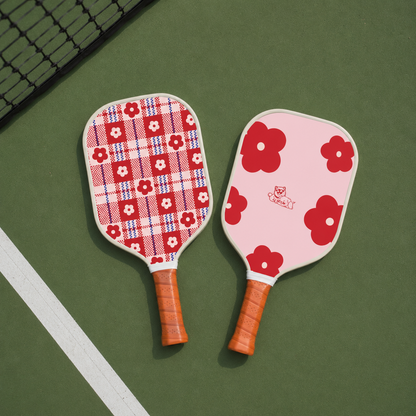 Deux raquettes de pickleball originales au sol, l'une rose à grosses fleurs rouges et l'autre à motif écossais rouge et blanc.