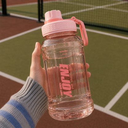 Main tenant une gourde de sport rose transparente de 1000ml au-dessus d'un terrain de pickleball.