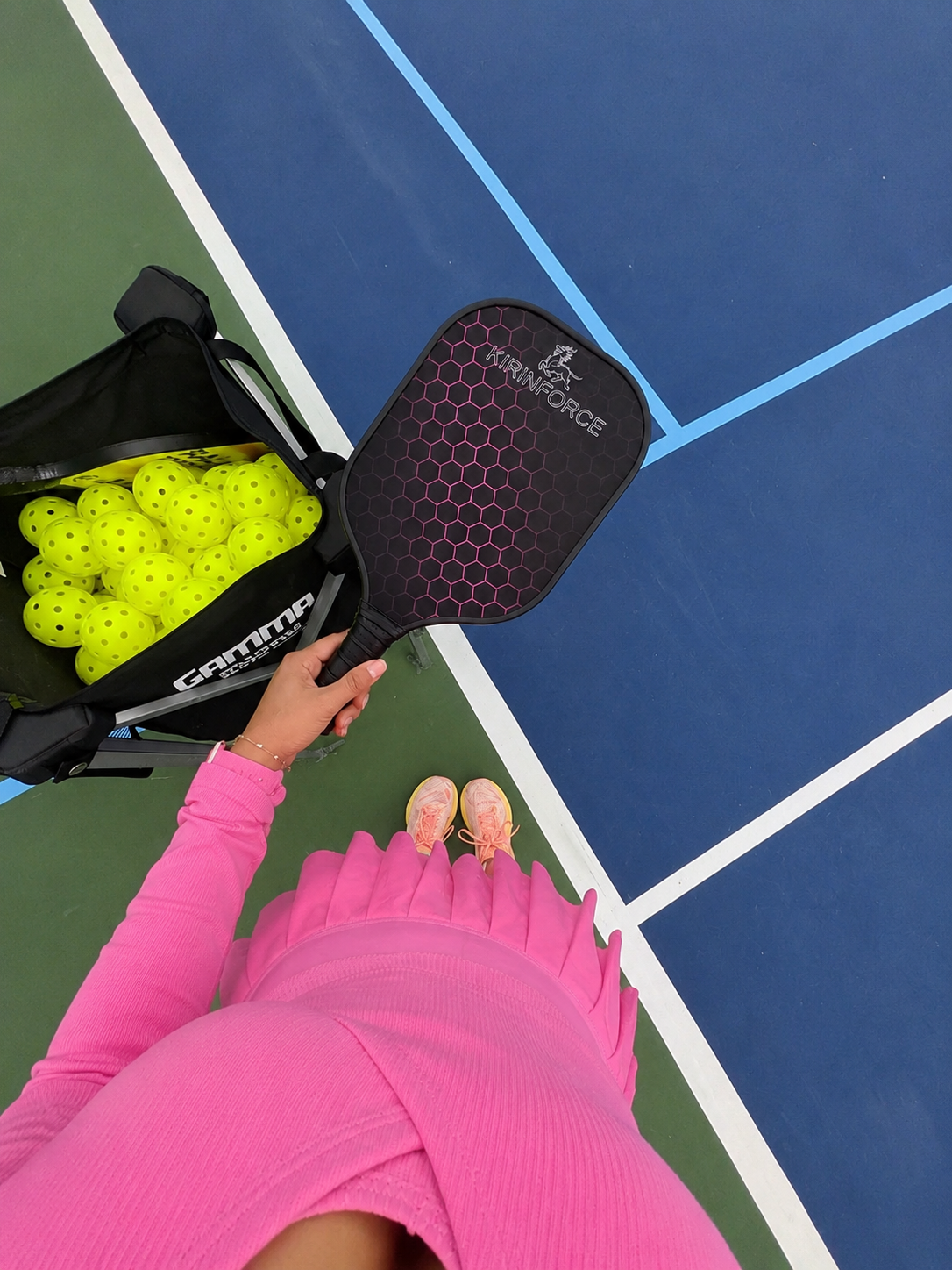 Vue POV d'une joueuse en tenue de sport rose tenant une raquette Kirinforce à motif hexagonal rose près d'un caddie de balles.