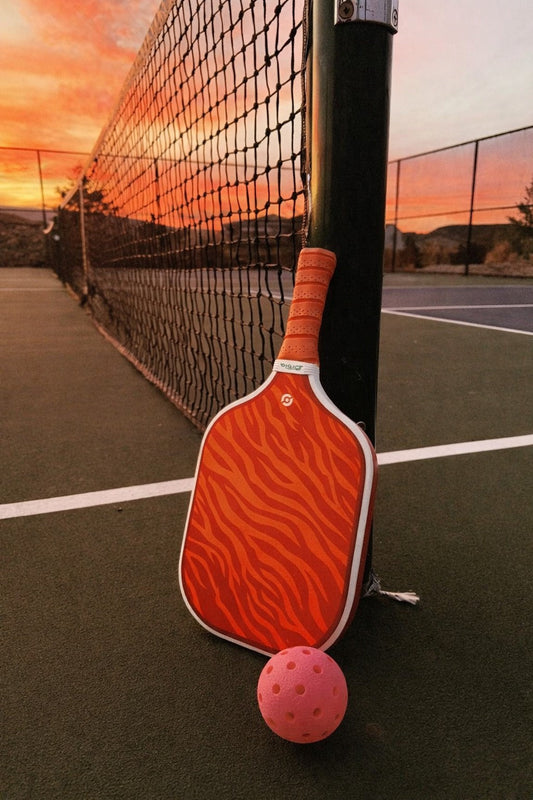 Raquette flamboyante Ember de Smashline posée sur un terrain de pickleball avec un soleil couchant